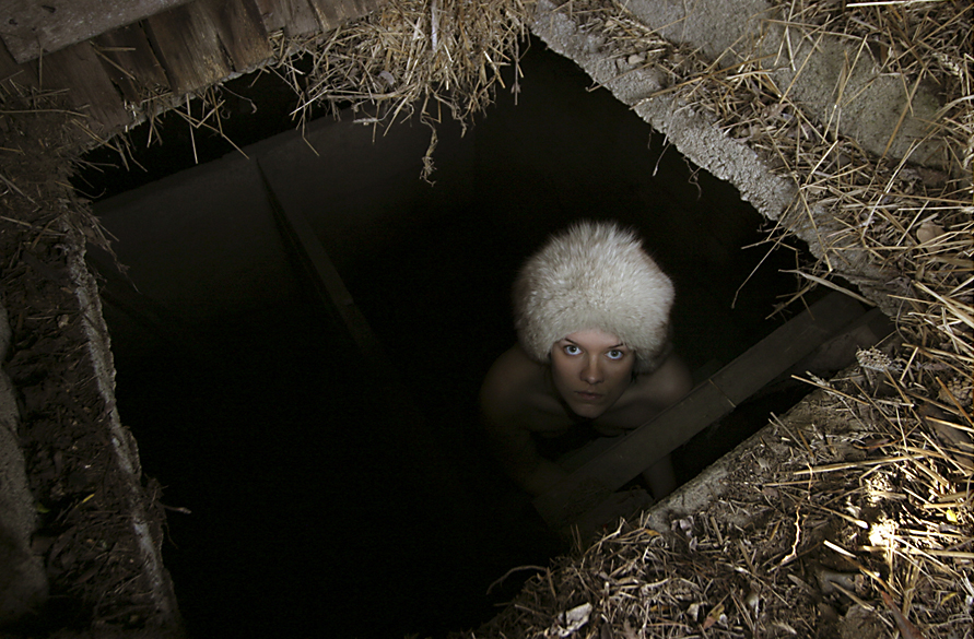 Photograph of a nude woman in a badger fur hat staring at the camera from a hole leading to a cellar.